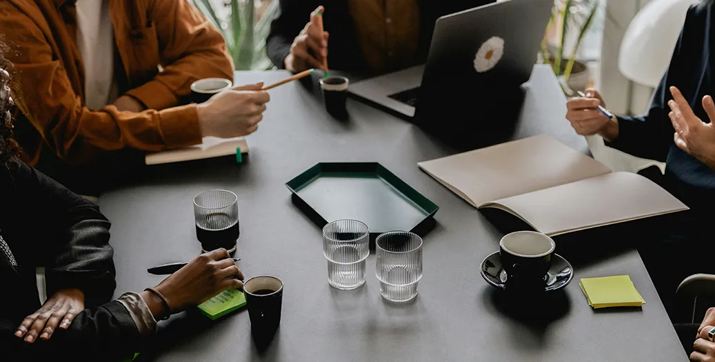 Reunião de trabalho em volta de uma mesa, com notebook, cadernos e café